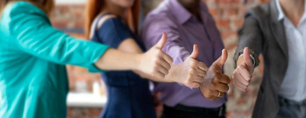 4 people stood next to each other holding out their right arms and making a thumbs up, all facing the right or the camera, their hands being clear while everything else is blurred