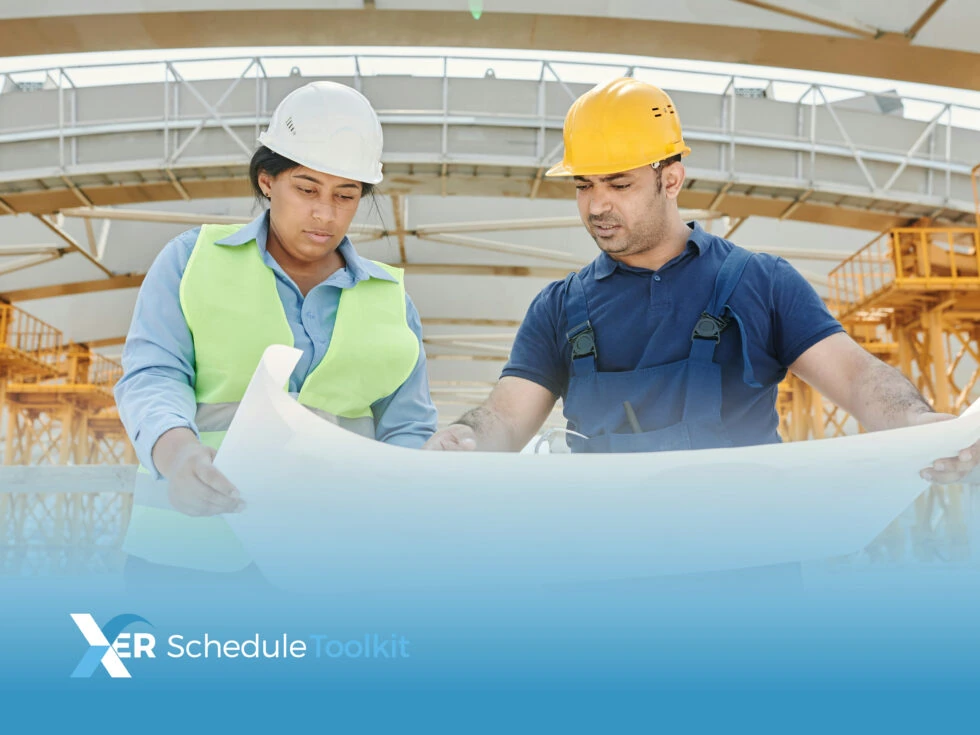 A man and woman in hard hats and work gear looking and pointing at a large piece of paper while stood on a site