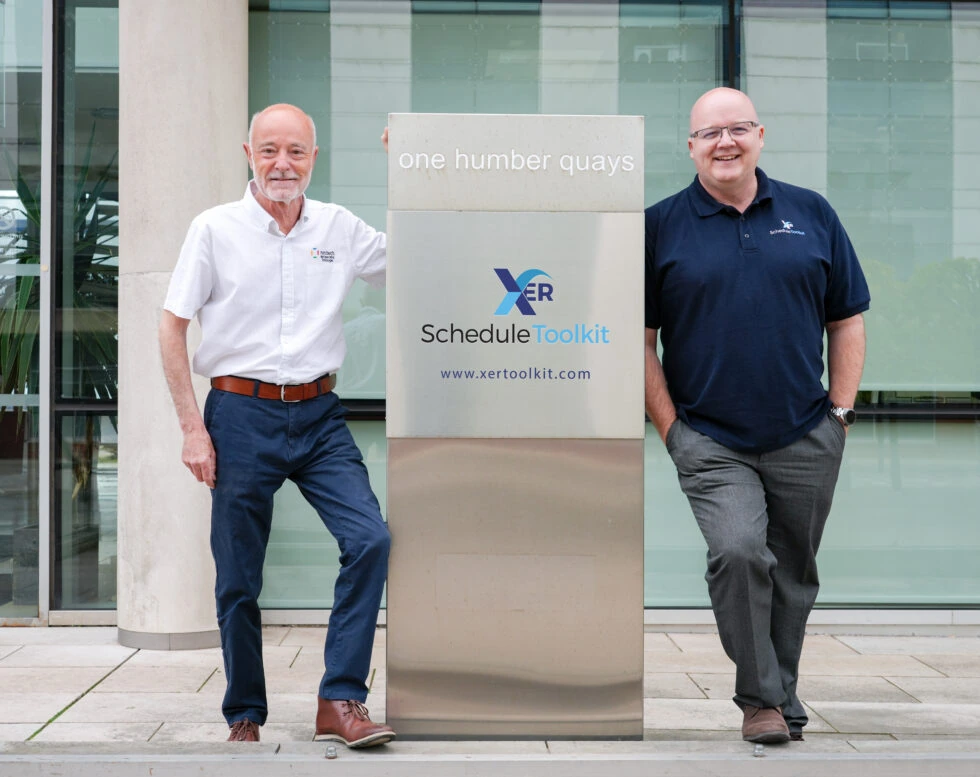 Chris Grsvenor and Tolis Chatzisymeon stood out front of the Spencer Group building, at the sign on the front with a plaque that states XER Schedule Toolkit with the link for the site below it