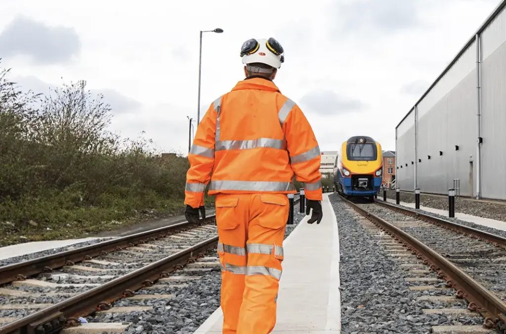 Man in hi-vis jacket, hi-vis trousers, and a hard hat walking between two train tracks